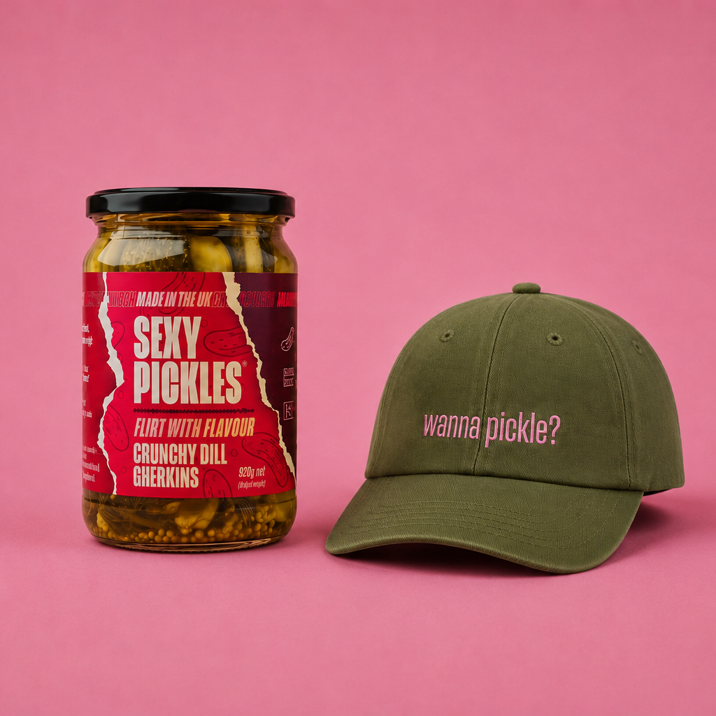A glass jar of 'Sexy Pickles' with a red label next to an olive green baseball cap that reads 'wanna pickle?' on a solid pink background.