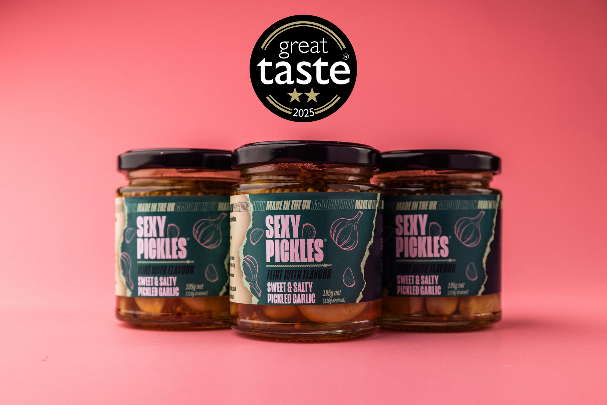 Three jars of 'Sexy Pickles' pickled garlic with a 'Great Taste 2025' award on a pink background